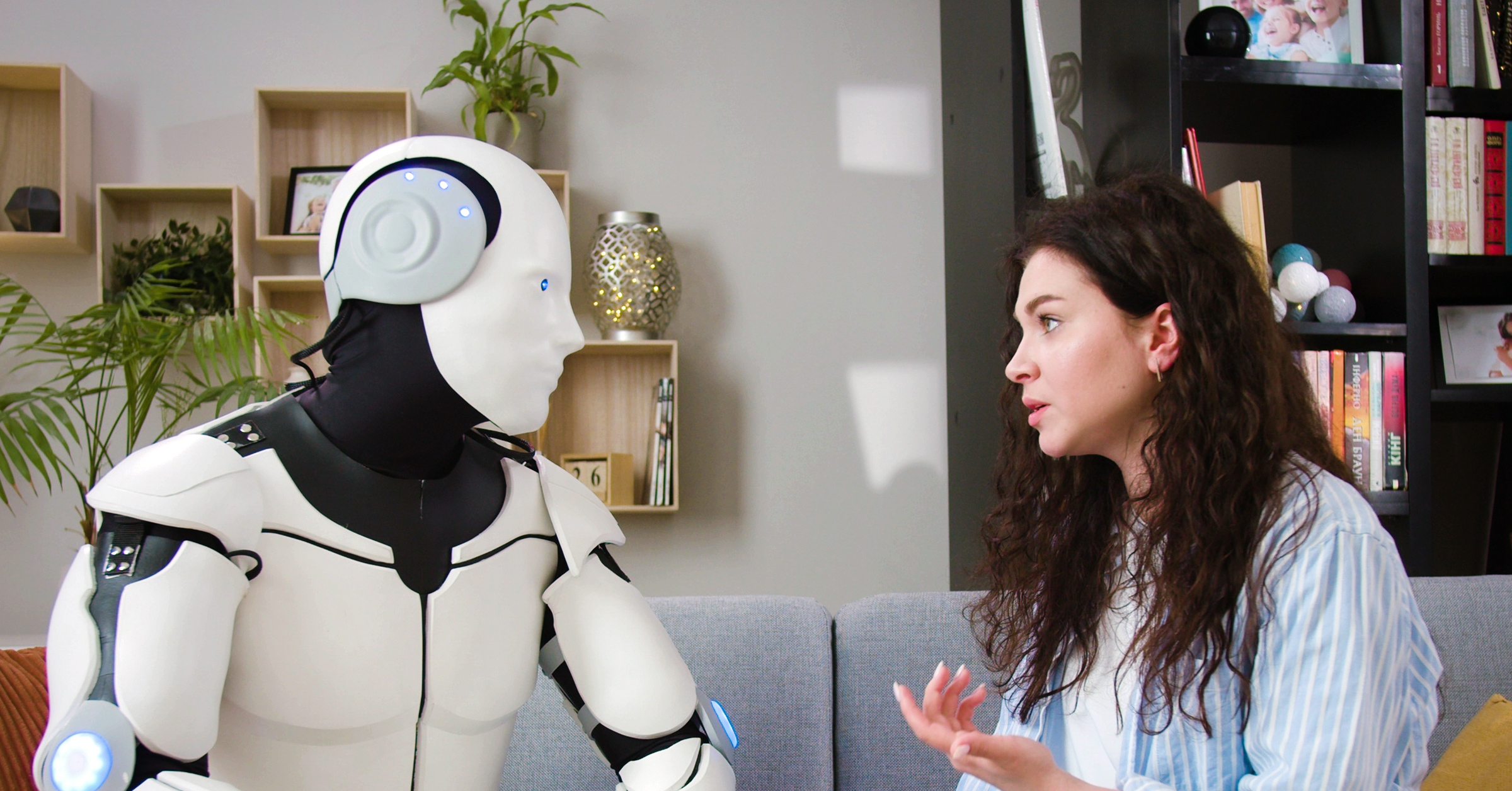 A woman with curly hair, wearing a white and blue shirt, is sitting on a grey couch in a living room, having an engaging conversation about customer storytelling with an AI humanoid robot. The room has shelves with decorative items, books, and a plant. The robot has a white and black exterior with blue lights.