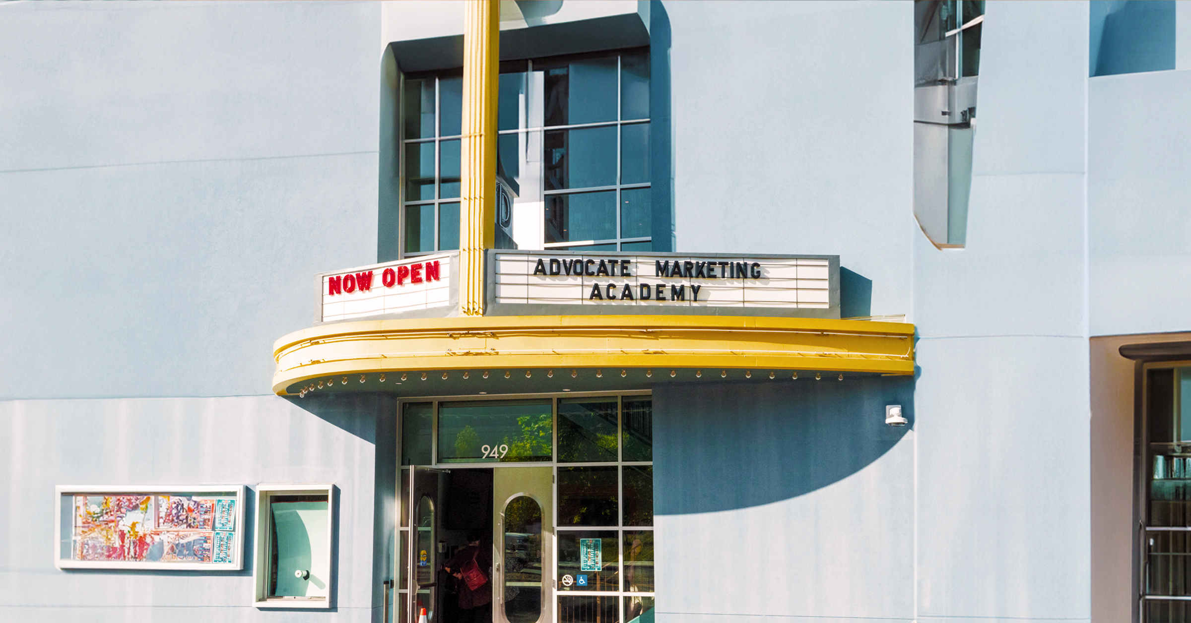 A light blue building with a curved yellow marquee displaying "NOW OPEN: ADVOCATE MARKETING ACADEMY - AMA USA". The entrance below has glass doors and a street number "949" above. There’s a small window display to the left and second-story windows above the marquee, highlighting its inviting facade.