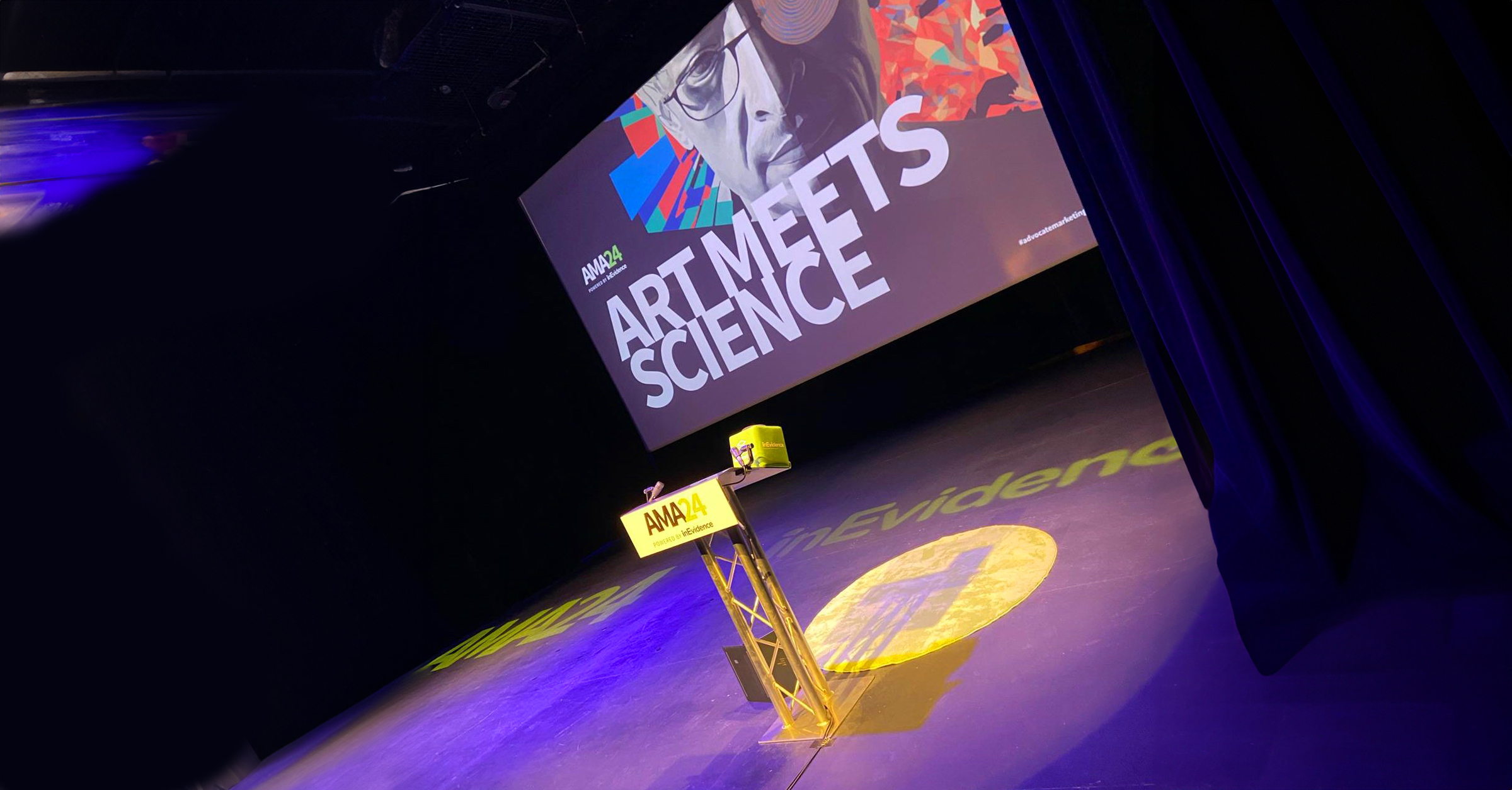 A stage with dark curtains and a large screen displaying the text "ART MEETS SCIENCE" in bold letters, with colorful abstract graphics and a stylized face. A podium with a yellow RA&M logo is positioned in front of the screen, with blue and green lighting enhancing the artistic scene.