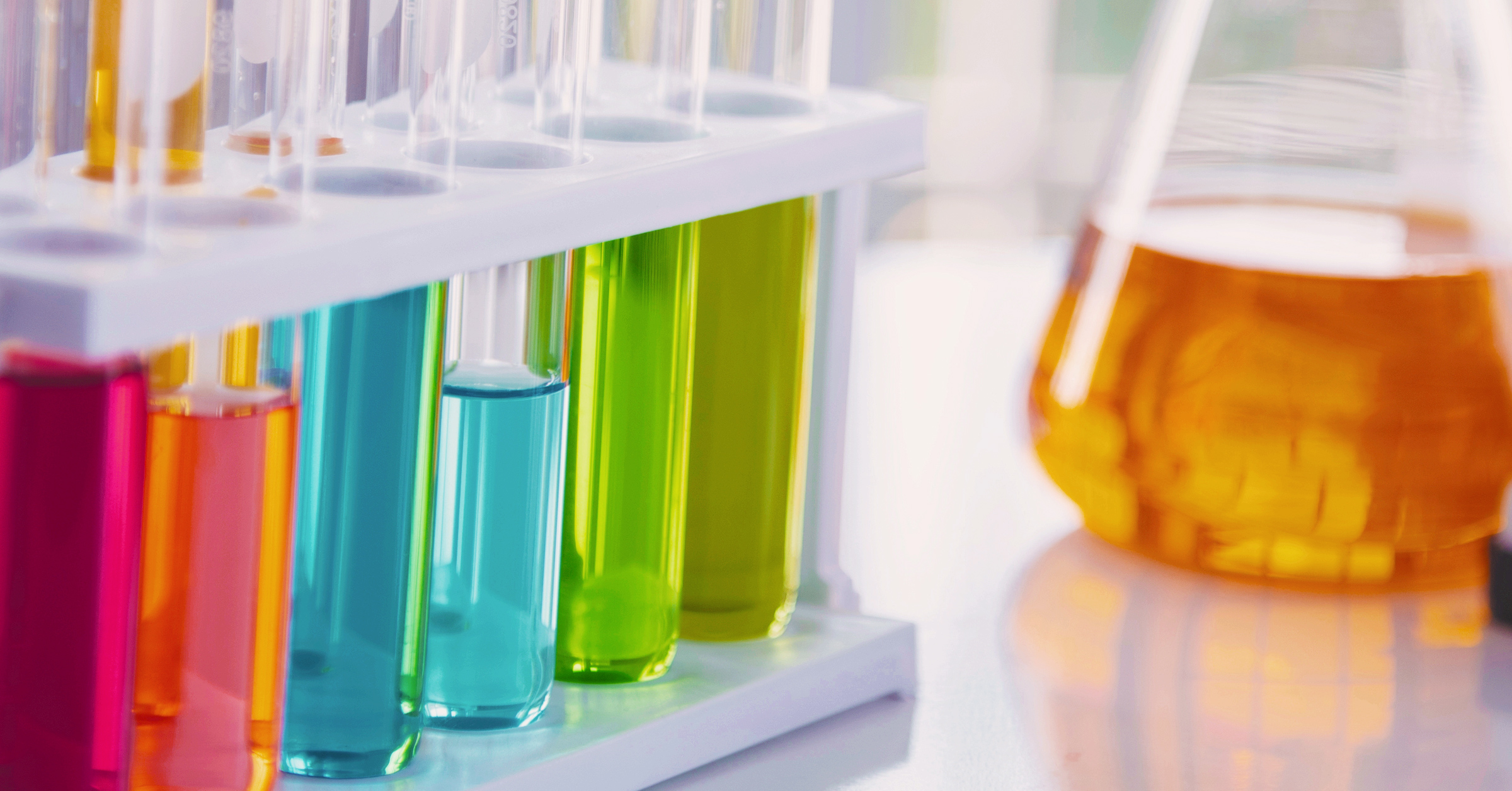 A collection of test tubes filled with colorful liquids, including red, blue, green, and yellow, arranged in a white test tube rack. An Erlenmeyer flask containing an orange liquid is positioned to the right. The setup is on a laboratory bench, reflecting the innovation trends highlighted in the 2024 Global Survey.