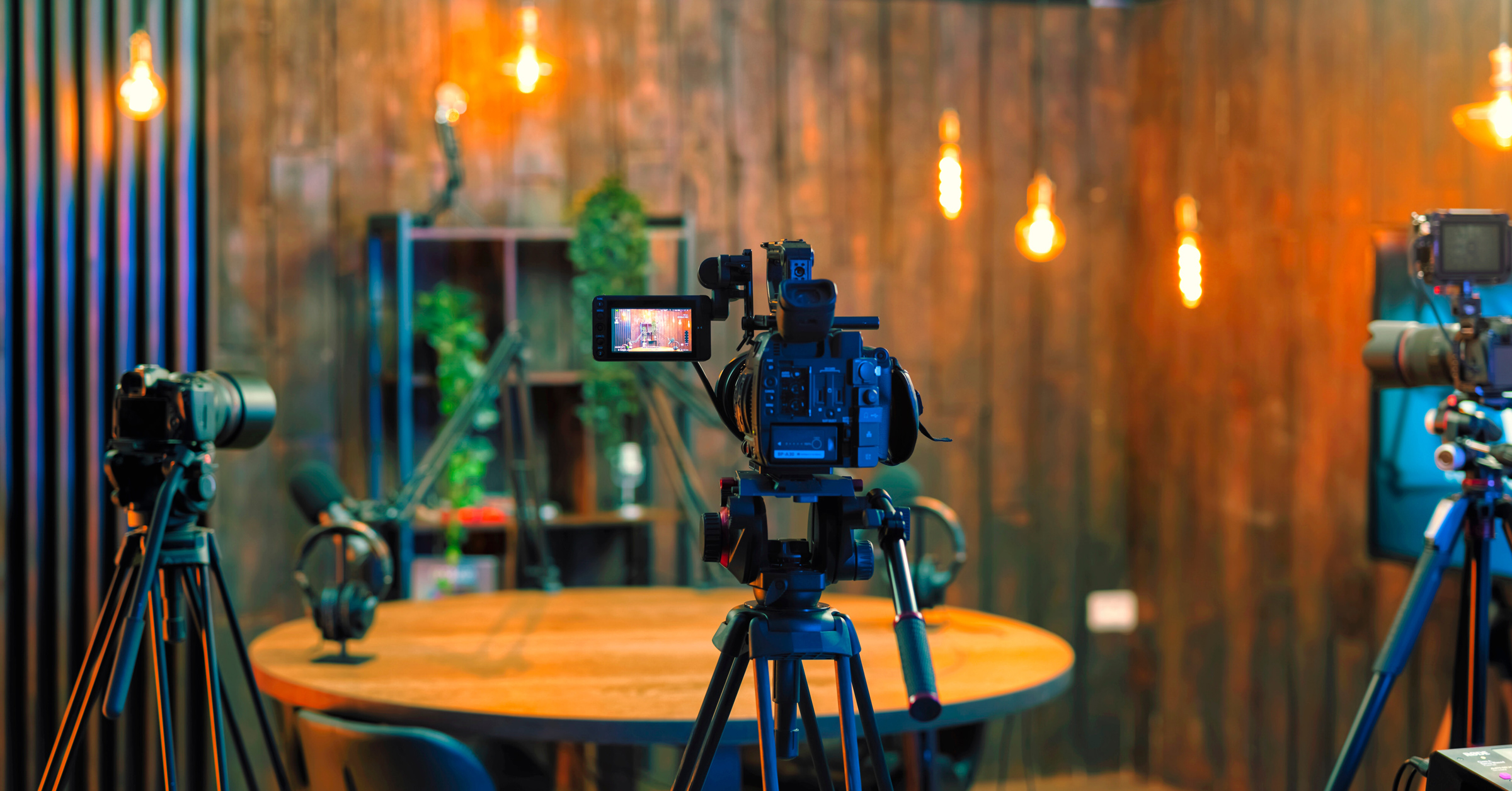 A cozy podcast or recording studio with wooden walls, warm Edison bulb lighting, and several cameras set up around a circular table with microphones and headphones. The scene appears ready for recording or a live broadcast, perfect for discussing topics like customer advocacy.